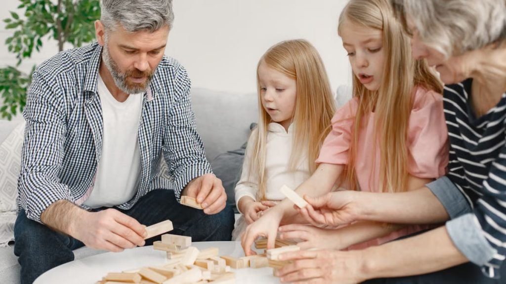 Four family members of different ages engage with toy blocks on a table indoors.