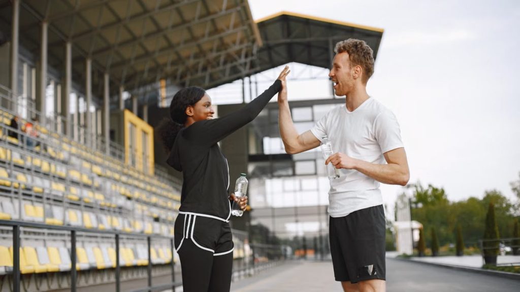 Two friends in activewear high-fiving with water bottles at an outdoor stadium, celebrating fitness.